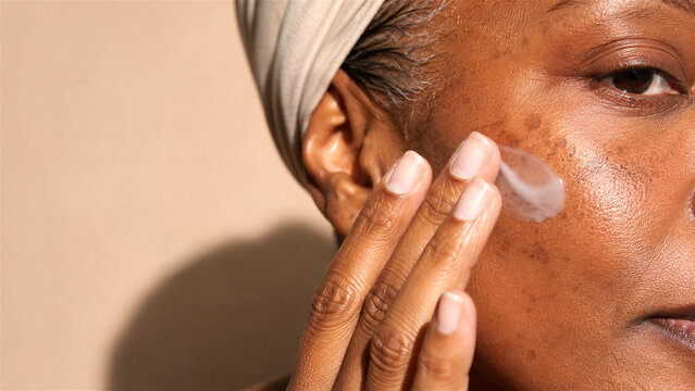 mature African American woman applying skincare serum  to cheek. Closeup beauty routine promoting self care, healthy aging, wellness, and dermatology treatment with copy space