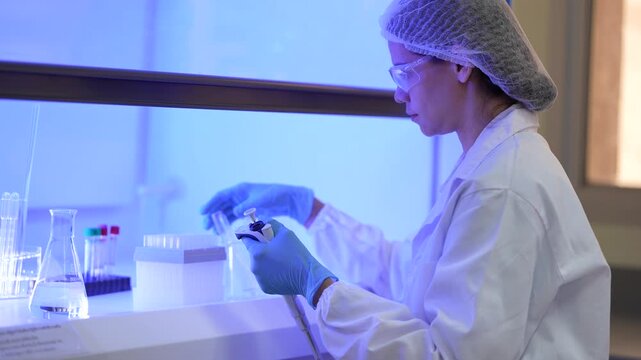 A young woman scientist in a white lab coat, hairnet and clear safety goggles collecting a tube from the rack then drawing sample up into the pipette in a modern medical research laboratory.