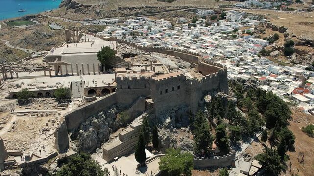 Stunning aerial drone footage flying over the ancient Acropolis of Lindos in Rhodes, Greece. The footage reveals the historic ruins perched above the picturesque whitewashed village of Lindos