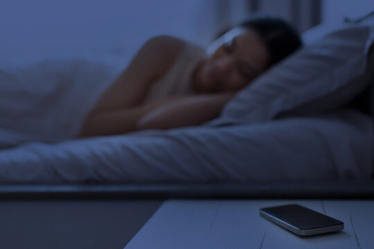 Young Black woman sleeping in dark bedroom at night while smartphone resting on bedside table, illustrating bedtime, rest, nighttime routine, and digital lifestyle at home