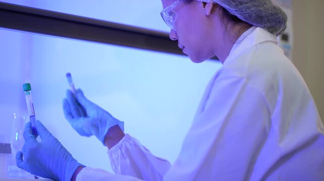 A woman researcher in a white lab coat, hairnet and clear safety goggles rotating a capped sample tube in her fingers then reaching to set it aside at a professional laboratory workstation.