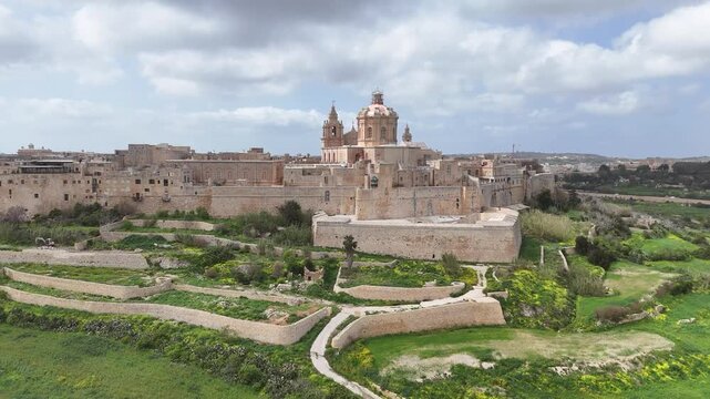 mdina ditch gardens bastion walls aerial view pathways ramparts moat honey limestone st pauls cathedral dome skyline rabat malta silent city medieval citadel hilltop fortress drone footage farmland