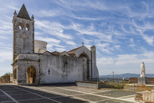 View of the historic Capela da Rainha Santa Isabel with its stone tower and tiled roof, set in a paved square under a blue sky with wispy clouds in Estremoz, Evora District, Portugal.