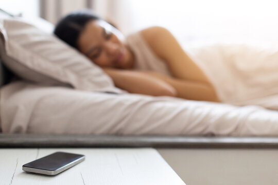 Young Black woman sleeping peacefully in bed while her smartphone resting on bedside table in the foreground, illustrating rest, morning routine, and digital lifestyle at home