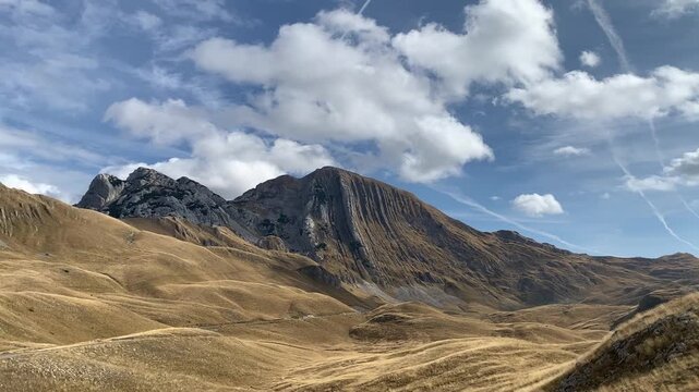 A wide golden grassy valley stretches toward dramatic mountain peaks under a cloudy blue sky in Durmitor Montenegro, scenic travel landscape, outdoor nature exploration, tranquil trip.
