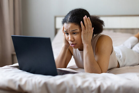 Young Black woman lying on her bed using laptop and holding her head in shock, appearing stressed or overwhelmed by online work, study, or a computer problem at home