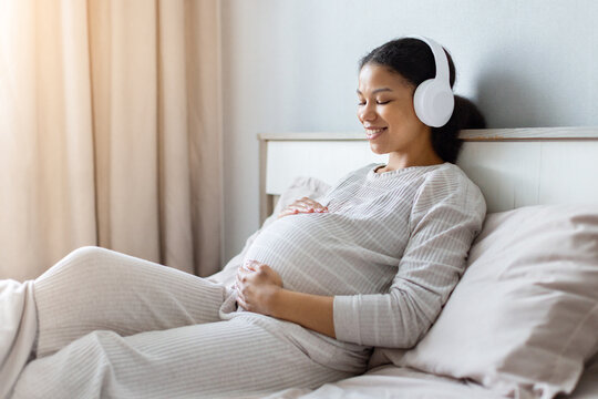 Smiling pregnant Black woman wearing wireless headphones while relaxing on her bed and holding her baby bump, enjoying music and a peaceful prenatal moment at home