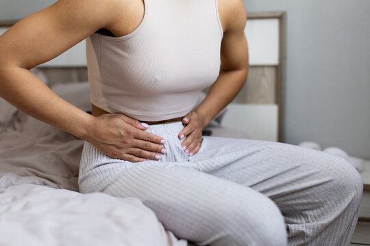 Closeup of young Black woman sitting on bed and holding her lower abdomen, experiencing stomach pain, cramps, or pelvic discomfort during a quiet moment at home