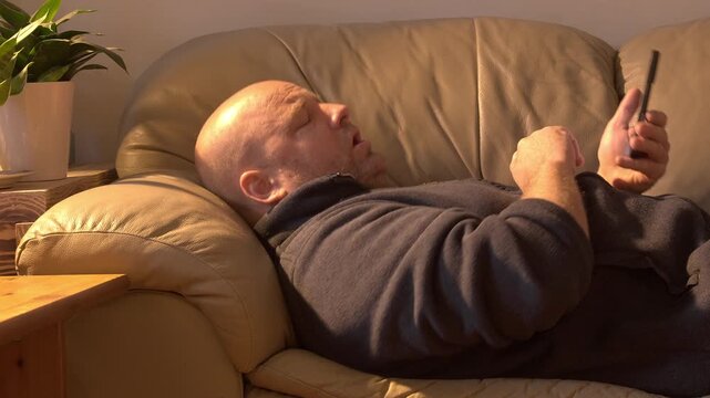 Middle-aged white man laying comfortably on a tan coloured leather sofa, doom scrolling on the cellphone, sneezes, and continued to scroll through the phone.