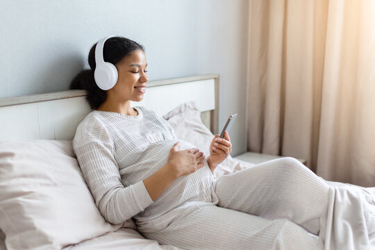 Smiling pregnant Black woman relaxing on her bed wearing wireless headphones, holding her baby bump and using smartphone during a peaceful moment at home