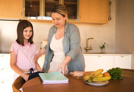 Woman and girl interacting in modern kitchen, preparing for meal with fresh fruits and vegetables on wooden table, bright and inviting atmosphere