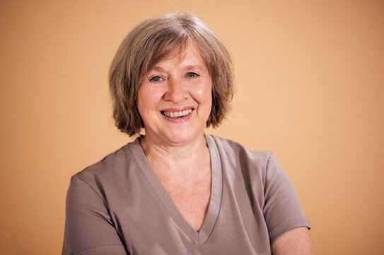 Senior woman with short gray hair smiles warmly while wearing a beige shirt against a soft, neutral background in a studio setting