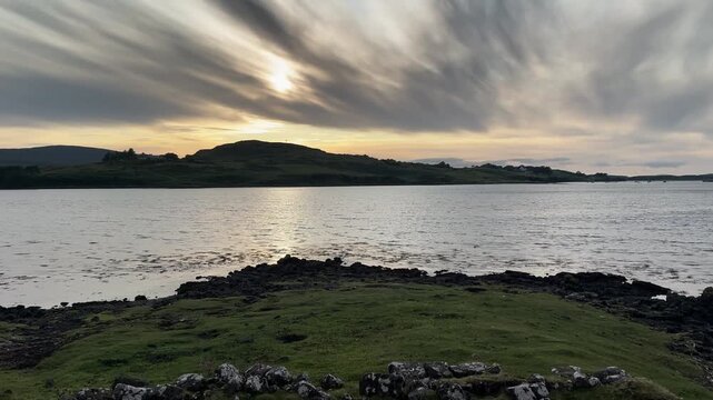 Dramatic sunset sky with fast moving clouds over a coastal loch in Scotland, rocky shoreline with green grass, moody atmospheric landscape.