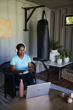 Woman sitting in converted container studio using laptop and microphone with punching bag nearby
