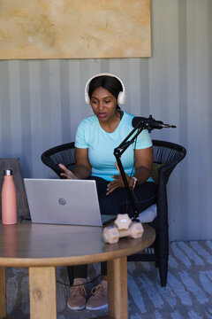 African woman in blue top and white headphones speaking into boom mic in studio using laptop