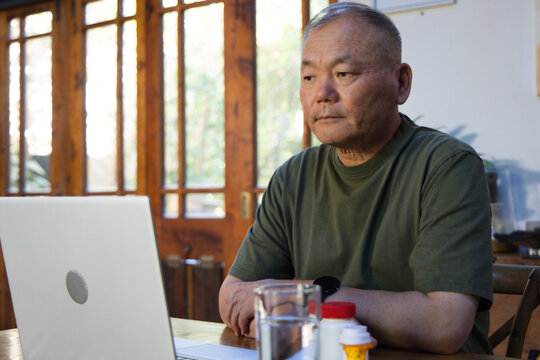Senior Asian man sitting at table by glass doors, olive-tee, viewing laptop with pills, copy space