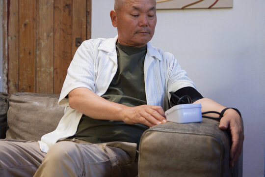Senior Asian man sitting at home on gray sofa wearing olive T-shirt checking blood pressure cuff