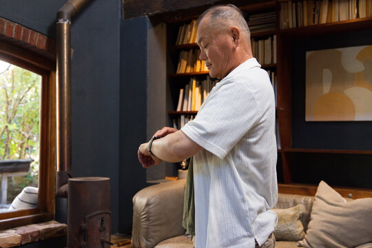 Senior Asian man standing by window in study, checking wristwatch, wearing short-sleeve shirt