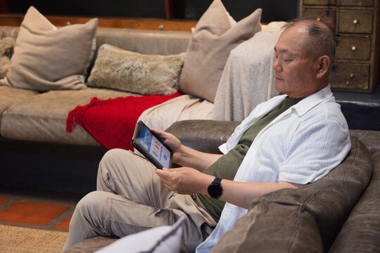 Senior man reclining on leather sofa in living room holding tablet wearing white shirt, copy space