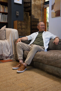 Senior Asian man sitting relaxed on brown sofa at home wearing olive T-shirt and beige chinos