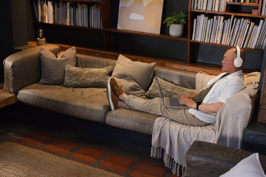 Senior Asian man reclining on leather sofa at home, using laptop, wearing headphones, copy space