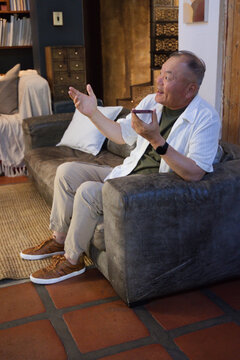 Senior Asian man sitting on dark gray sofa in living room, holding smartphone and gesturing