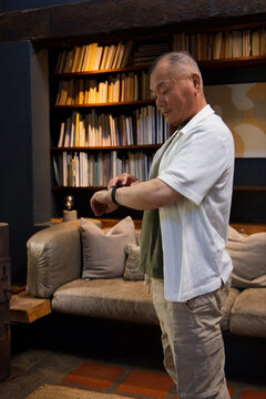 Senior Asian man checking wristwatch while standing in reading room with wooden bookcase and sofa