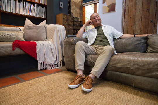 Senior Asian man sitting on brown leather sofa leaning back, looking left in living room