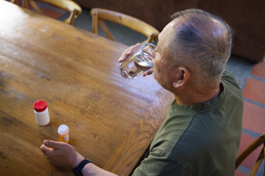 Senior Asian man sitting at table drinking from glass, pill bottles beside, olive shirt, copy space