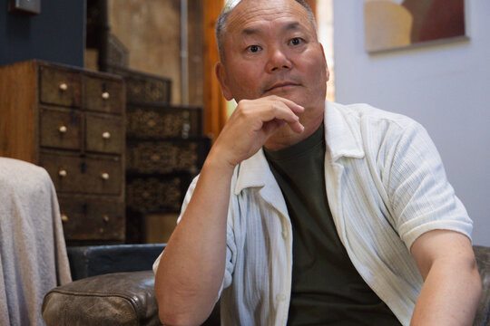 Senior Asian man sitting on leather, wearing striped shirt over green tee, looking at abstract art