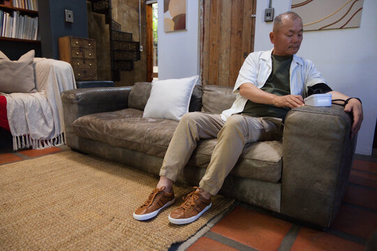 Senior Asian man checking blood pressure in living room on right sofa with cuff, copy space