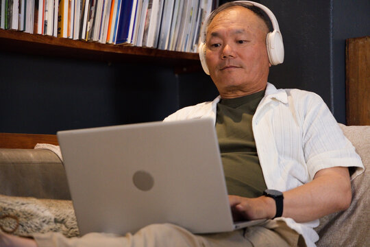 Senior Asian man sitting on cushioned sofa in living room using silver laptop and white headphones