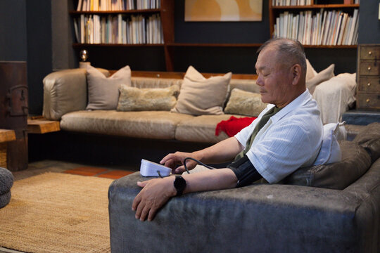 Senior Asian man sitting in armchair at home checking blood pressure monitor on lap, copy space