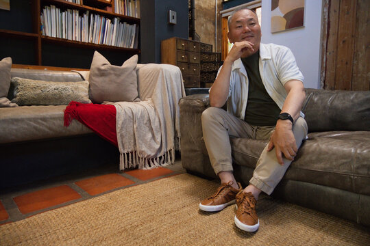 Senior Asian man sitting on right leather sofa in lounge, resting chin, showing watch, copy space