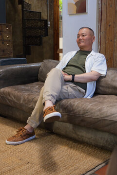 Senior Asian man sitting on worn brown leather sofa in rustic lounge, legs crossed, watching