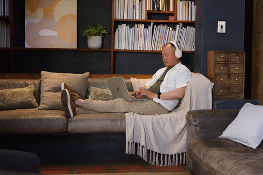 Senior man reclining on leather sofa in cozy living room, using laptop and wearing headphones