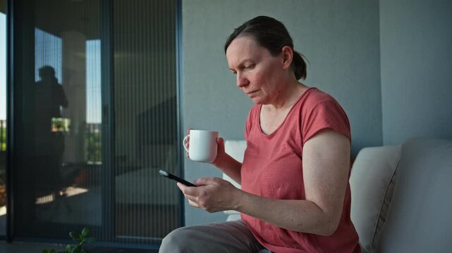 Middle-aged woman holding coffee mug, checking smartphone on balcony sofa, while husband&rsquo;s reflection appears in glass door using his phone. 4K.