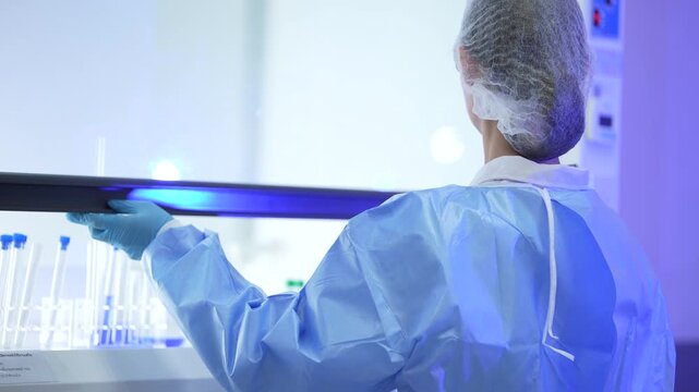A woman researcher in a white lab coat, hairnet and clear safety goggles seen from behind she lifts a tube into the cabinet, sets it, reaches again at a professional laboratory workstation.