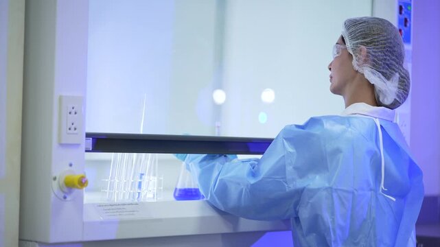 A female laboratory technician in a white lab coat, hairnet and clear safety goggles seated at the fume hood she sweeps a hand across the tube rack in a modern medical research laboratory.