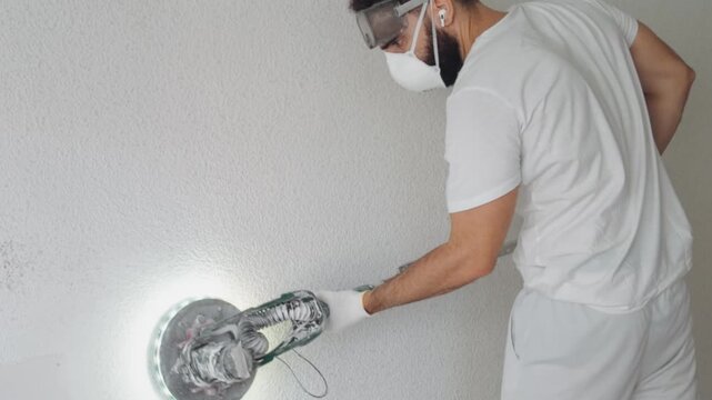 Male worker wearing safety mask and glasses uses electric sander with glowing light to smooth white plaster wall during interior home renovation and diy house construction project