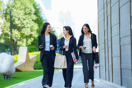 A team of three Hispanic businesswomen in matching dark suits, laughing openly as they walk, walk three abreast along a sunlit avenue carrying tote bags, folders and phones.