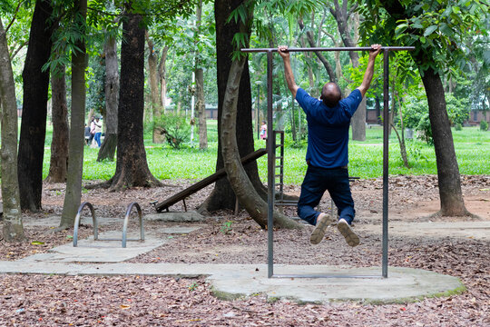 Dhaka, Bangladesh - 26 May 2024: View of a man performing pull-ups on a metal exercise bar in Ramna Park, surrounded by lush trees and greenery.