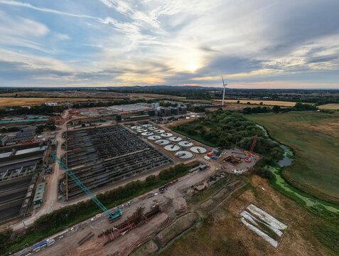 Aerial view of the wastewater treatment plant featuring circular sedimentation tanks, construction cranes, and a wind turbine in Wanlip, England, United Kingdom.