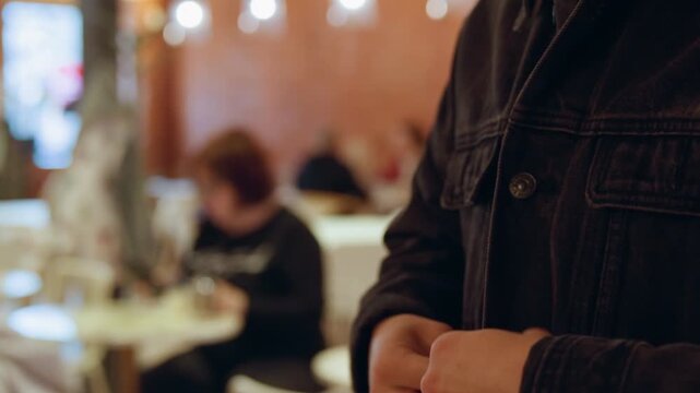 Close up of man buttoning his dark denim jacket in indoor setting with blurred background activity. focus remains on hands and lower torso capturing steady movement as man fastens each cuff