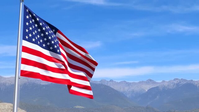 Slow motion pan of american flag waving on flagpole against mountain sky with clear daylight and distant peaks