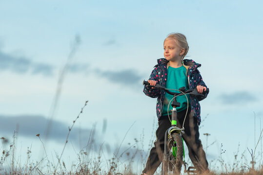 Blonde child standing on a bicycle in an outdoor field, dressed in a jacket and looking thoughtfully towards the horizon, symbolizing discovery, growth, and the journey of childhood
