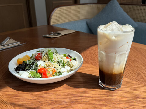 Colorful Mixed Salad With Tomatoes, Greens, Cheese And Orange Slice Sits Beside Tall Iced Coffee On Wooden Table In Bright Restaurant Seating Area, Suggesting Relaxed Lunch, Menu, Hospitality