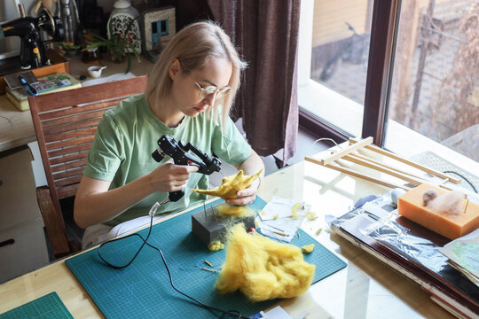 Young Woman With Glasses Works Carefully On Wool Sculpture At Home Craft Table, Holding Craft Tool And Shaping Yellow Fiber Material Near Window Light, With Felt Supplies, Mat, Foam Block