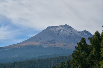 Majestic Popocatépetl seen from Paso de Cortés, rising above a dense green forest as clouds drift across a blue sky. The scene blends height, nature, and serenity, capturing one of Puebla © Omar