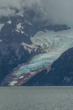 Majestic glacier flowing down steep dark mountain peaks into cold ocean water under cloudy overcast sky